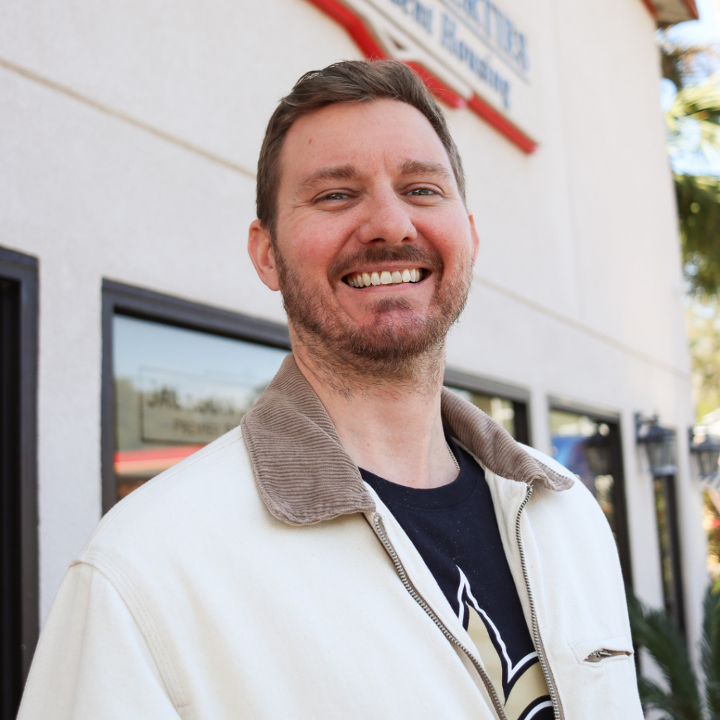Portrait photo of Brian Jackson in front of the Jackson Properties office in Tallahassee, Florida. He is smiling at the camera wearing a black t-shirt with a white and tan jacket over it.