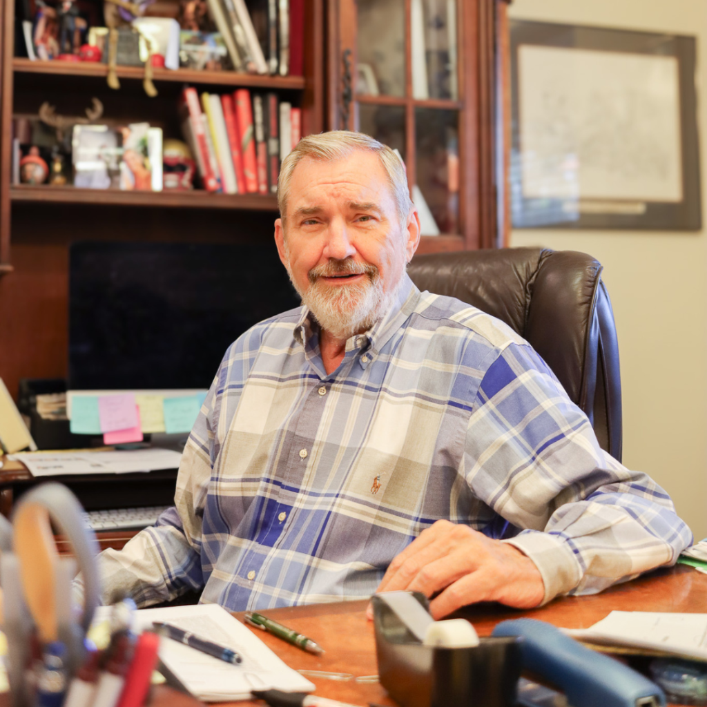 Portrait photo of Erwin Jackson at his desk at Jackson Properties in Tallahassee, Florida. He has on a white, grey, and blue plaid shirt and is facing the camera with a smile. In the background you can see a bookshelf with books in it.