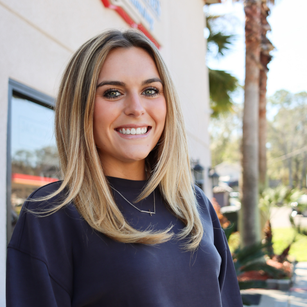 Portrait photo of Blair Hurst in front of the Jackson Properties in Tallahassee, Florida. Blair is smiling at the camera wearing a navy blue crewneck sweater and a necklace. Her hair is blonde and straightened.